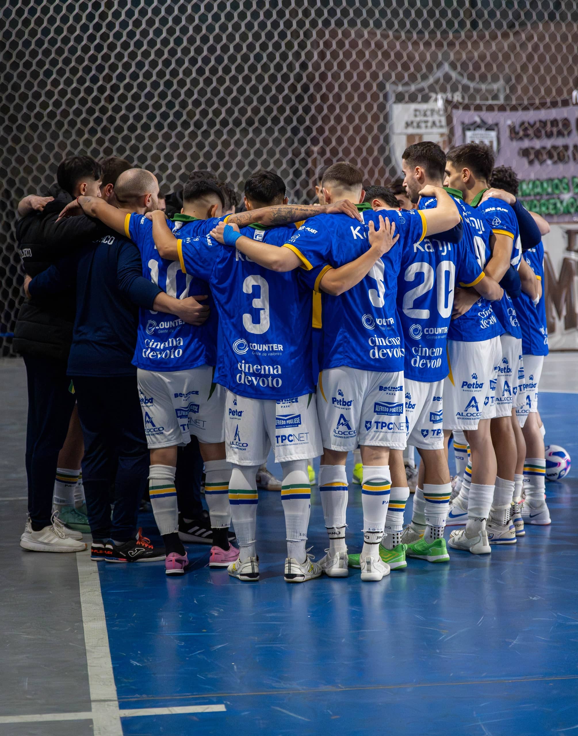 Un plantel completo abrazado en círculo, de espaldas, con camisetas azules y shorts blancos, sobre el piso azul de una cancha cubierta.