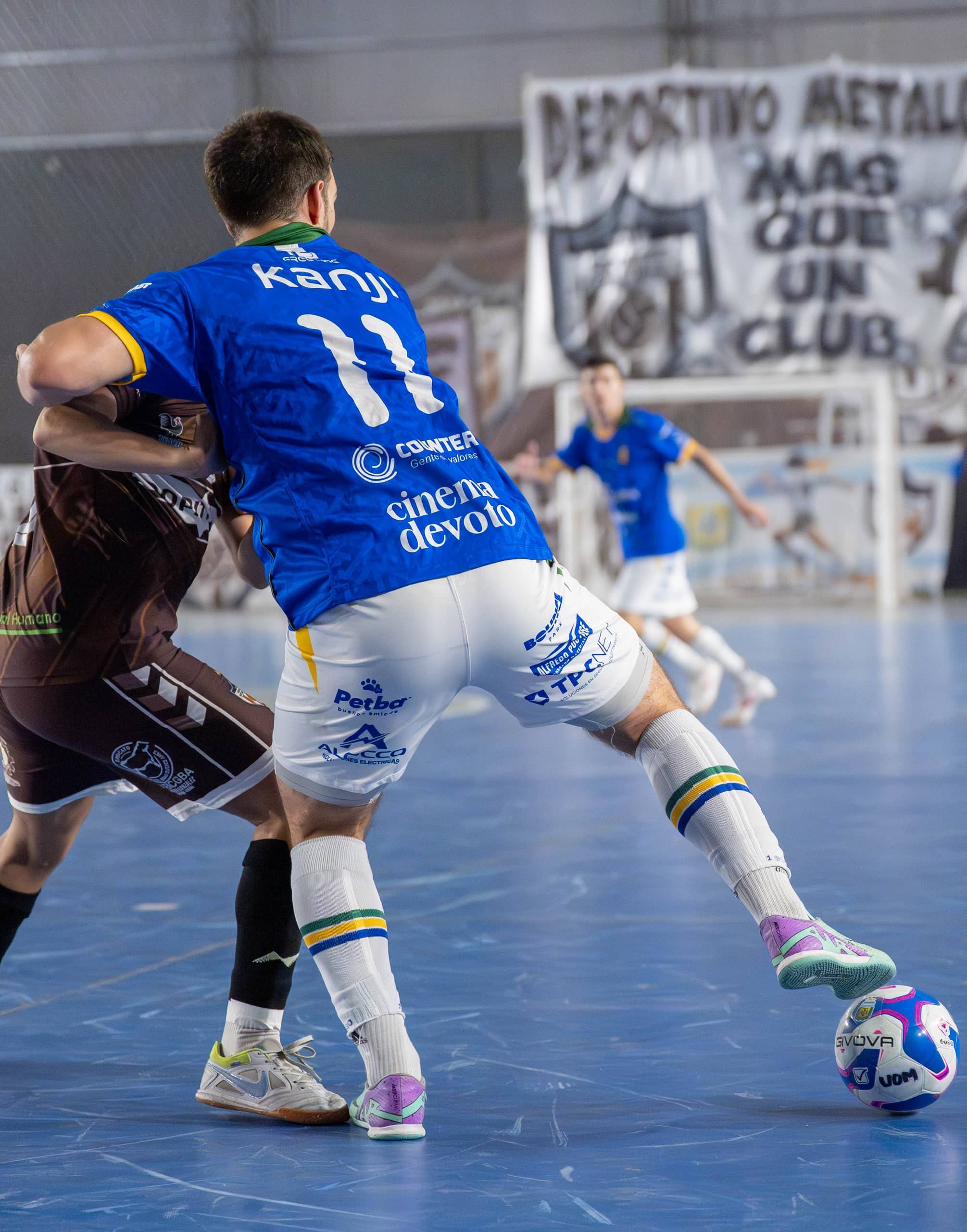 Un jugador de camiseta azul y short blanco protege la pelota con el cuerpo frente a un rival de conjunto marrón oscuro, sobre el piso azul de una cancha cubierta; al fondo, un arco y una bandera colgada.