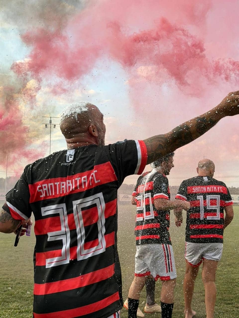 Jugadores de espaldas festejando en una cancha al aire libre, con camisetas a rayas rojas y negras y el dorsal a la vista; el aire está teñido de humo rojo y hay agua salpicando.