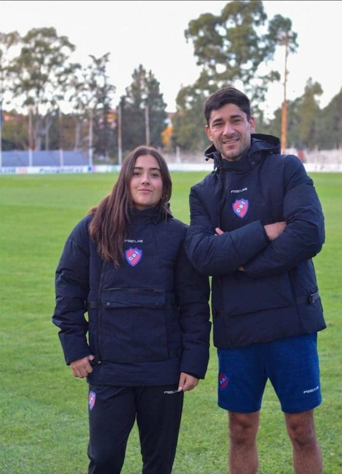 Una chica y un hombre de pie sobre el césped de una cancha, los dos con camperas azul marino con el escudo de una institución en el pecho.