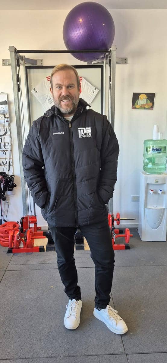 Un hombre de pie en un gimnasio con una campera negra de la marca y una pelota de pilates detrás.