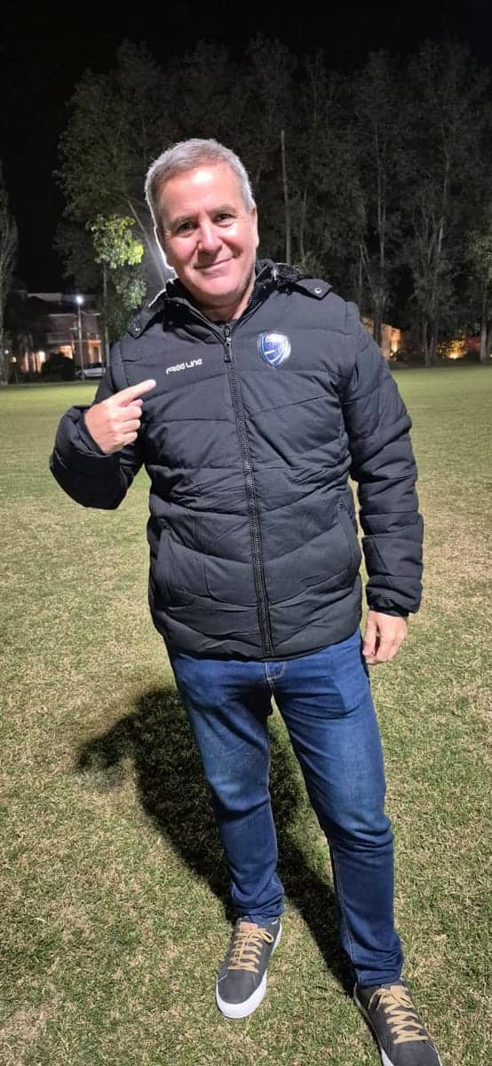 Un hombre señala el escudo de su campera azul oscuro, de noche, al costado de una cancha iluminada.