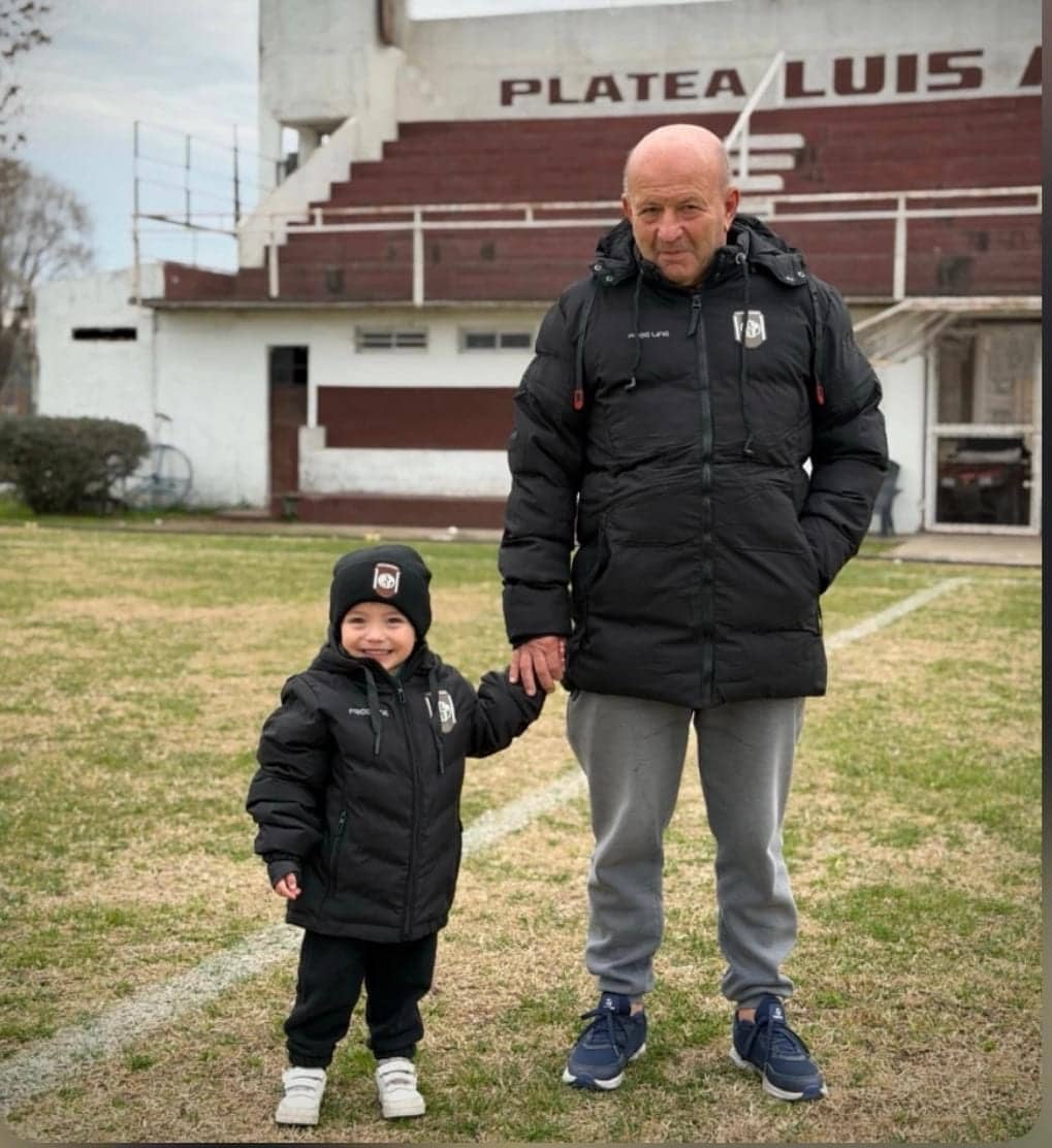 Un hombre mayor y un nene de la mano sobre el césped, los dos con camperas negras iguales; atrás, la platea de un club.