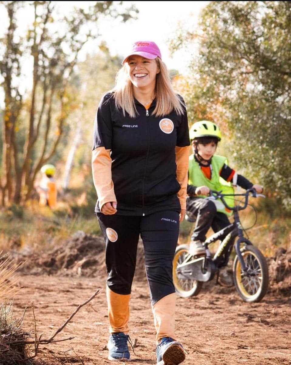 Una mujer camina por un sendero de tierra con una remera negra de la marca y una gorra rosa; detrás, un chico en bicicleta con casco.