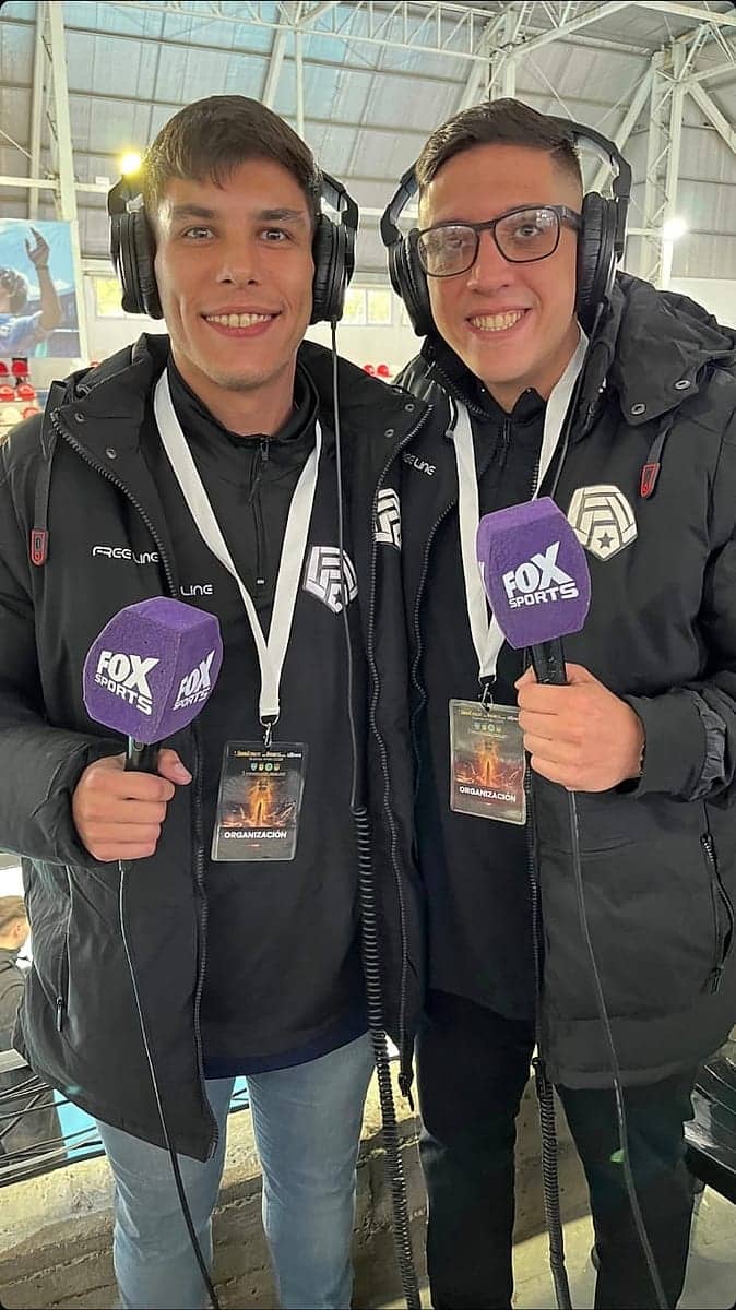 Dos personas con auriculares y micrófonos de mano, con camperas negras de la marca, en el borde de una cancha.